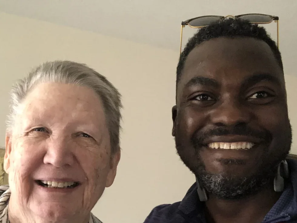 Pierre Beauge, BSN, RN, smiling beside an older client in her living room